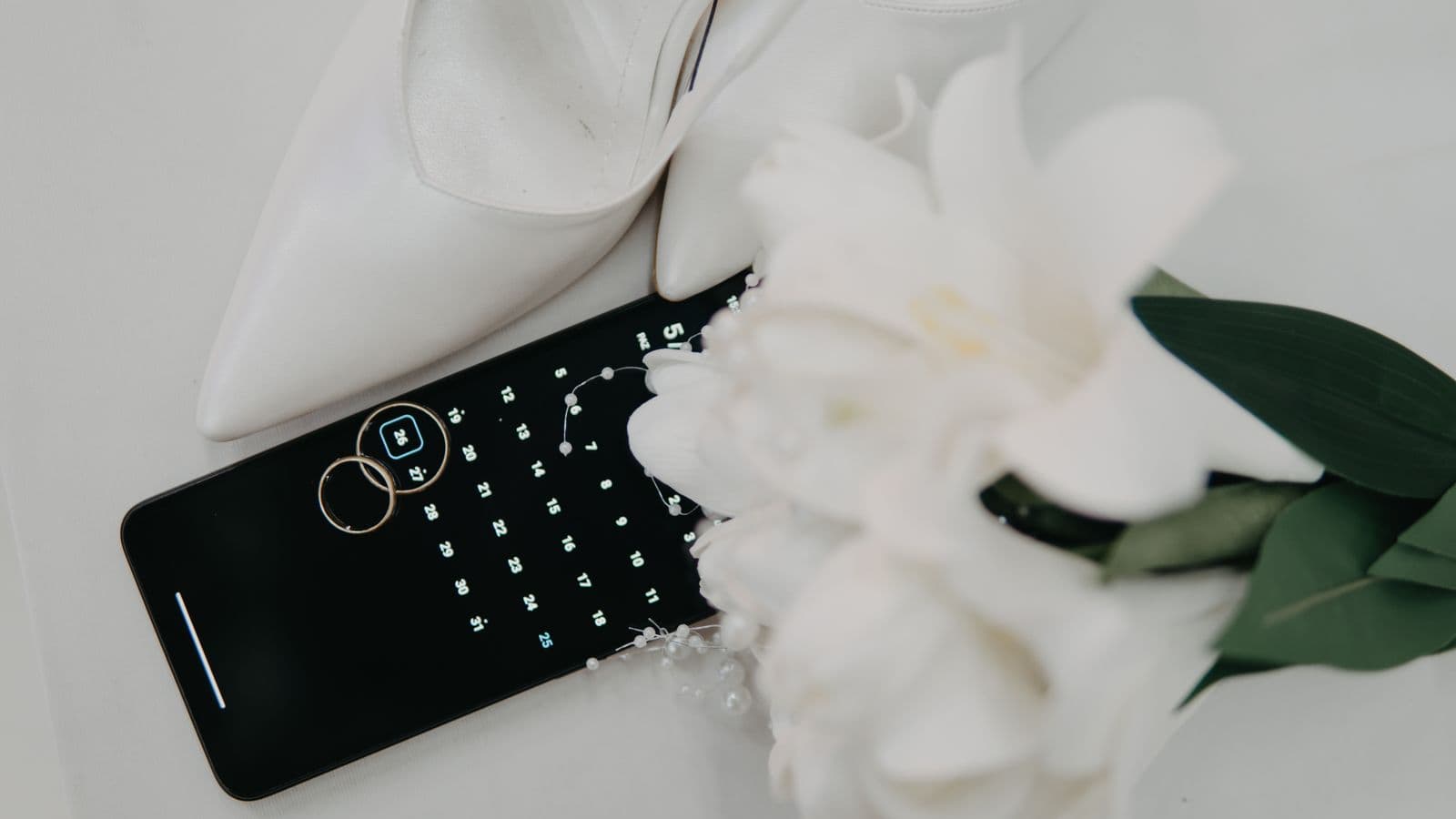 Phone calendar with a marked wedding date beside rings, bridal shoes, and a bouquet
